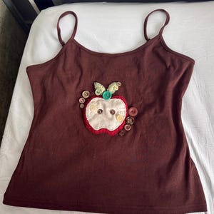 May include: A brown spaghetti strap tank top featuring a central fabric apple appliqué. The apple is constructed from cream and red fabric, decorated with various small buttons in shades of brown, cream, and green, including a prominent teal button.
