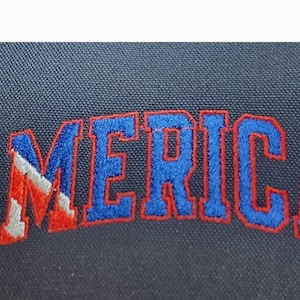 May include: Embroidered "AMERICA" in red and blue thread on a dark blue fabric background. The "A" is in the style of the American flag.