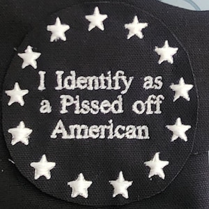 May include: A black fabric patch with a circle of white stars around the edge. The text "I Identify as a Pissed off American" is embroidered in white in the center of the patch.