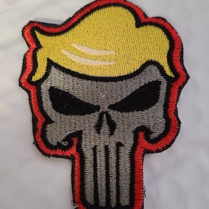 May include: A gray and black embroidered skull with a yellow hair patch, outlined in red. The skull is a popular symbol of the Punisher, a fictional character from Marvel Comics.