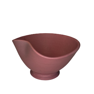 May include: A maroon-colored ceramic sauce bowl with a spout. The bowl has a rounded shape with a small, circular base. The spout is on the left side of the bowl. The bowl is empty and sits against a white background.