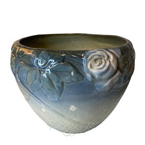 May include: A blue ceramic bowl with a floral design. The bowl features embossed roses and leaves around the rim. The interior is a darker blue-green, with a lighter blue gradient towards the bottom.