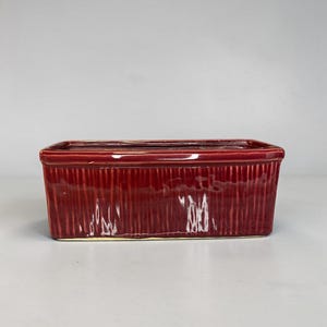 May include: A rectangular, burgundy-colored ceramic baking dish. The dish has a ribbed texture on the sides and a smooth, glossy finish. The interior is a lighter shade of burgundy.