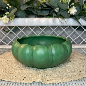 May include: A green ceramic bowl with a pumpkin-like design, resting on a beige crocheted doily. The bowl has a textured surface and a scalloped rim. The background features white wicker and greenery.
