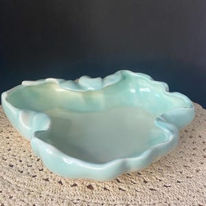 May include: A light blue ceramic bowl with a glossy finish and an irregular, wavy, ruffled rim. The bowl features a smooth, concave interior and a curved, organic shape that mimics the appearance of a shell or a cloud, with soft color gradients.