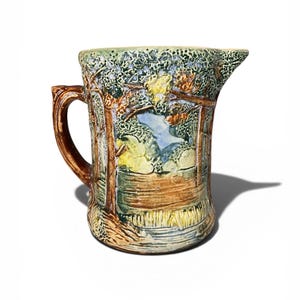 May include: A ceramic pitcher with a detailed landscape design. The pitcher features a brown handle and spout, with a scene of trees, water, and foliage in shades of green, blue, yellow, and brown. The overall style is rustic and artistic.