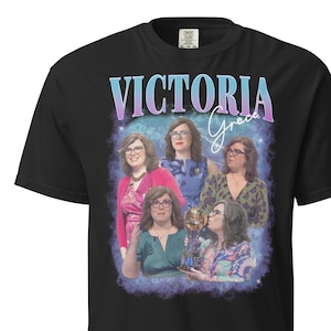 May include: Black t-shirt with a vintage-style graphic of Victoria Grace, featuring a group of women with a trophy and the text "Victoria Grace" in a pink and blue gradient.