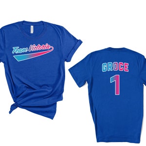 May include: Two royal blue t-shirts. One shirt has the text "Team Victoria" in a rainbow gradient arc. The other shirt has the text "GROCE" and the number "1" in a rainbow gradient.