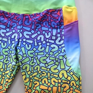 May include: Rainbow leggings with a colorful question mark and exclamation point pattern. The leggings have a yellow pocket on the right side.
