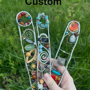 May include: Three custom-made wire bookmarks with various beads, stones, and charms. The bookmarks are silver in color and have a variety of colors and patterns. The word "Custom" is written in white letters on a green background above the bookmarks.