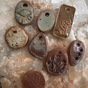 May include: A collection of seven small, irregular ceramic pendants with holes for stringing, featuring various glazes in brown, green, and grey tones, some with embossed floral or eye designs, displayed next to a copper-colored US one-cent coin.