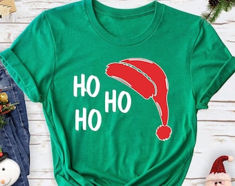 May include: A green t-shirt featuring the text "HO HO HO" and a red Santa hat illustration. The shirt is displayed on a white wooden surface, accompanied by blue jeans, wrapped gifts, and festive Christmas decorations.