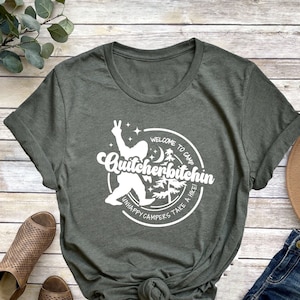 May include: A dark green t-shirt with a white graphic design featuring a cartoon figure with the text "Welcome to Camp Quitcherbitchin Unhappy Campers Take a Hike!"