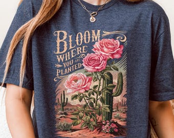 May include: Navy blue t-shirt featuring a desert scene with a cactus and pink roses. The shirt has the text "Bloom Where You Are Planted" in an ornate font. The design is printed on the front of the shirt.