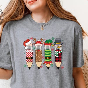 May include: A gray t-shirt featuring a festive pencil design. Four pencils are decorated with holiday themes: a Santa hat, reindeer antlers, an elf hat, and a top hat with a scarf. The pencils have red, brown, green, and plaid patterns.