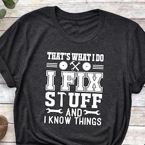 May include: Dark gray t-shirt with the text "THAT'S WHAT I DO I FIX STUFF AND I KNOW THINGS" in white. The shirt is styled with a brown hat, blue jeans, and brown sandals.