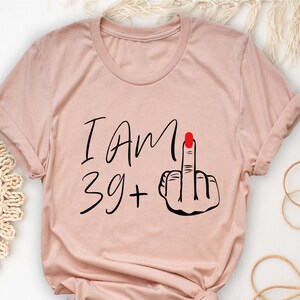 May include: A light pink t-shirt with the text "I Am 39 +" and a black and red illustration of a middle finger.