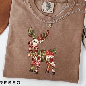 May include: Espresso-colored t-shirt featuring a festive reindeer design. The reindeer is adorned with a patchwork of red, green, and white patterns, including bows, poinsettias, and candy cane stripes. A silver necklace with a pendant is draped across the shirt.