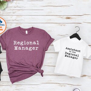 May include: A black onesie, a burgundy t-shirt, and a white t-shirt, each with text. The burgundy shirt reads "Regional Manager." The white shirt and black onesie read "Assistant to the Regional Manager."