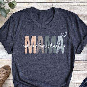 May include: A dark blue t-shirt with the word "MAMA" in colorful letters and the text "Love Michael" underneath. The shirt is on a wooden surface with a pair of brown sandals, a pair of blue jeans, and a brown hat.