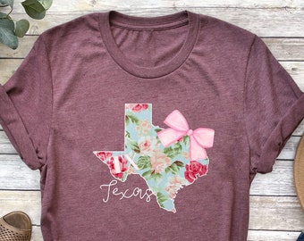 May include: A maroon t-shirt with a floral Texas outline and a pink bow. The state shape is filled with a floral pattern in pink, blue, and white. The word "Texas" is written in cursive below the state outline.