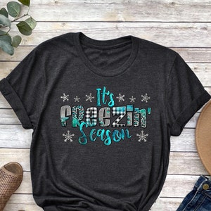May include: Dark gray t-shirt with the text "It's Freezin' Season" in turquoise, black and white plaid, and leopard print. The shirt is styled with a brown hat, blue jeans, and brown sandals.