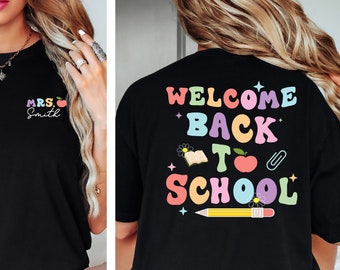 Puede incluir: Camiseta negra con un diseño arcoíris de "Welcome Back to School" en la espalda y "MRS. Smith" con una manzana roja en el frente.