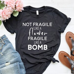 May include: Black t-shirt with the text "NOT FRAGILE LIKE A FLOWER FRAGILE LIKE A BOMB" in white. The shirt is paired with blue jeans, pink flowers, and brown sandals. The shirt is knotted at the waist.