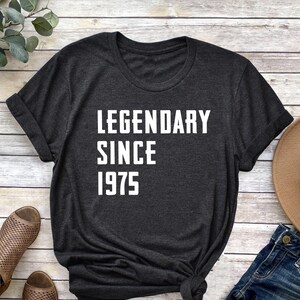 May include: A dark gray t-shirt with the text "LEGENDARY SINCE 1975" printed in white on the front. The shirt is laid flat on a wooden surface with a pair of brown sandals, a pair of blue jeans, and a brown hat.