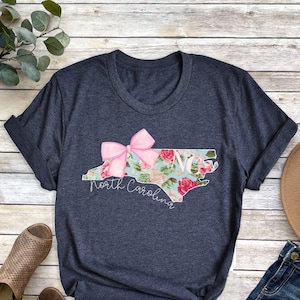 May include: A dark blue t-shirt featuring a floral design of the state of North Carolina with a pink bow on top. The text "North Carolina" is written below the state outline.