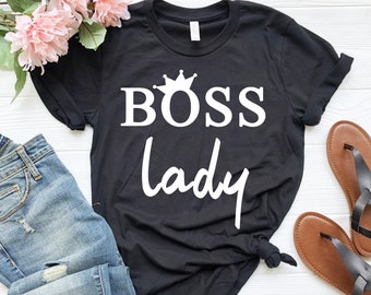 Pode incluir: Camiseta preta com as palavras "BOSS LADY" em branco, encimada por um gráfico de coroa. A camisa é combinada com jeans azuis, sandálias marrons e flores cor-de-rosa em uma superfície de madeira branca.