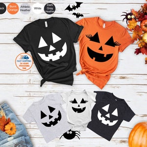 May include: A collection of Halloween-themed apparel, including black, orange, gray, and white t-shirts and a onesie. Each item features a jack-o'-lantern face design. The image also includes a pumpkin, decorative leaves, and a pair of jeans.