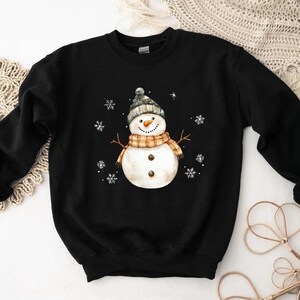 May include: Black sweatshirt featuring a watercolor snowman design. The snowman wears a gray and white knit hat, an orange and tan plaid scarf, and has twig arms. White snowflakes are scattered around the snowman. A cozy winter-themed apparel item.
