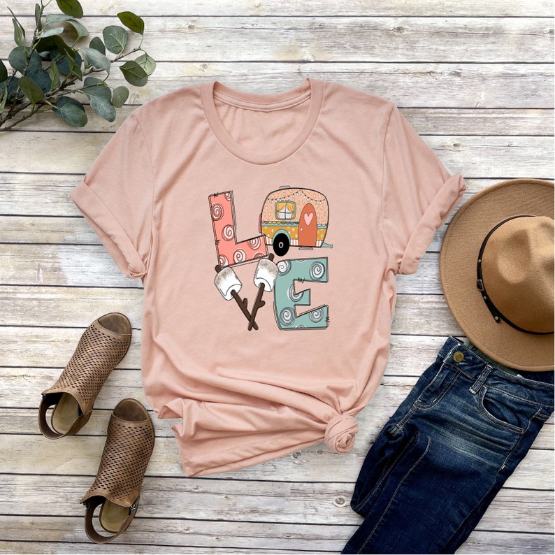May include: A pink t-shirt with a graphic design of the word "LOVE" with a camper van inside the "O". The letters are decorated with swirls and patterns. There are also two marshmallows on sticks in front of the "O".