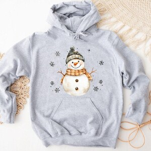 May include: A light gray hooded sweatshirt featuring a watercolor illustration of a snowman. The snowman wears a black and gray hat, an orange and brown scarf, and has twig arms. Black snowflakes are scattered around the snowman.