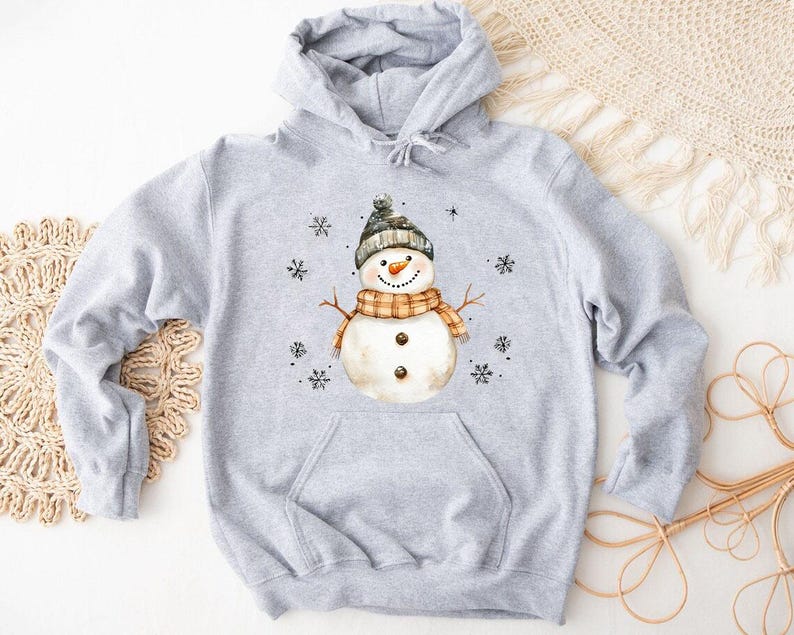 May include: A light gray hooded sweatshirt featuring a watercolor illustration of a snowman. The snowman wears a black and gray hat, an orange and brown scarf, and has twig arms. Black snowflakes are scattered around the snowman.