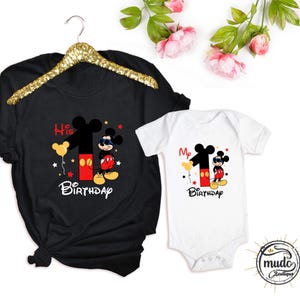 Op de afbeelding: Zwart T-shirt en witte romper met een Mickey Mouse-ontwerp voor een eerste verjaardag. Het ontwerp bevat het nummer 1, Mickey Mouse, ballonnen en de woorden "His Birthday" en "My Birthday".