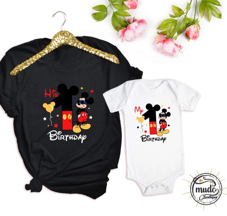 Op de afbeelding: Zwart T-shirt en witte romper met een Mickey Mouse-ontwerp voor een eerste verjaardag. Het ontwerp bevat het nummer 1, Mickey Mouse, ballonnen en de woorden "His Birthday" en "My Birthday".
