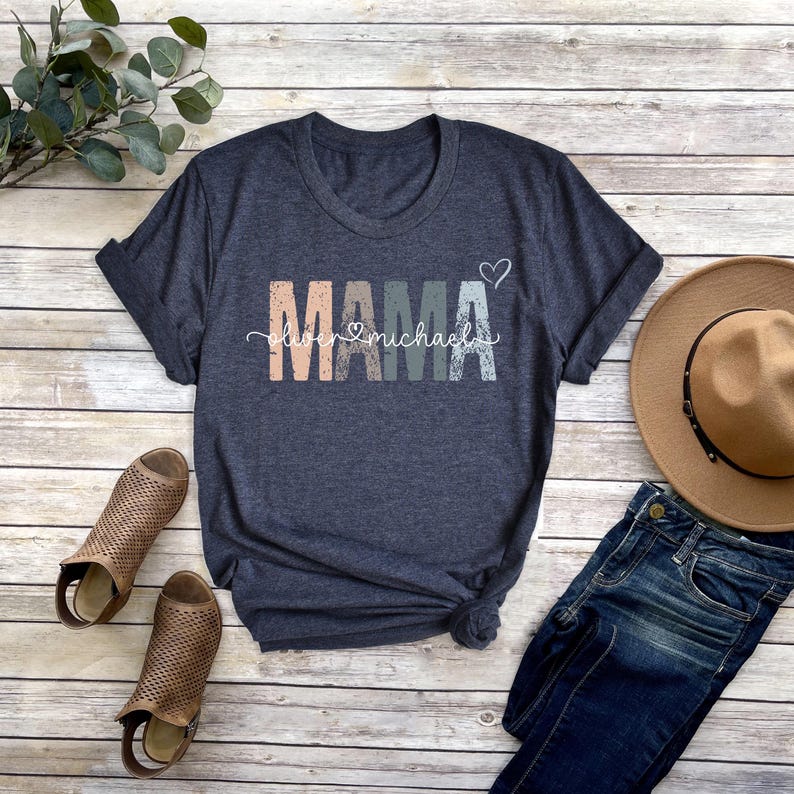 May include: A dark blue t-shirt with the word "MAMA" in colorful letters and the text "Love Michael" underneath. The shirt is on a wooden surface with a pair of brown sandals, a pair of blue jeans, and a brown hat.