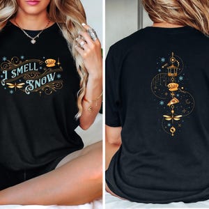 May include: Black t-shirt with the text "I Smell Snow" in a decorative font, with dragonfly and snowflake accents. The back of the shirt features a gold design with a lantern, pizza slice, and dragonfly.