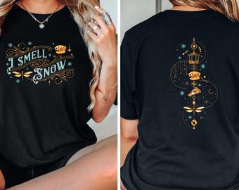 May include: Black t-shirt featuring the text "I Smell Snow" in a decorative font, with dragonfly and snowflake accents. The back of the shirt has a gold design with a lantern, pizza slice, and dragonfly.
