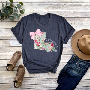 May include: A dark blue t-shirt with a floral print of the state of Louisiana. The print includes a pink bow and the word "Louisiana".