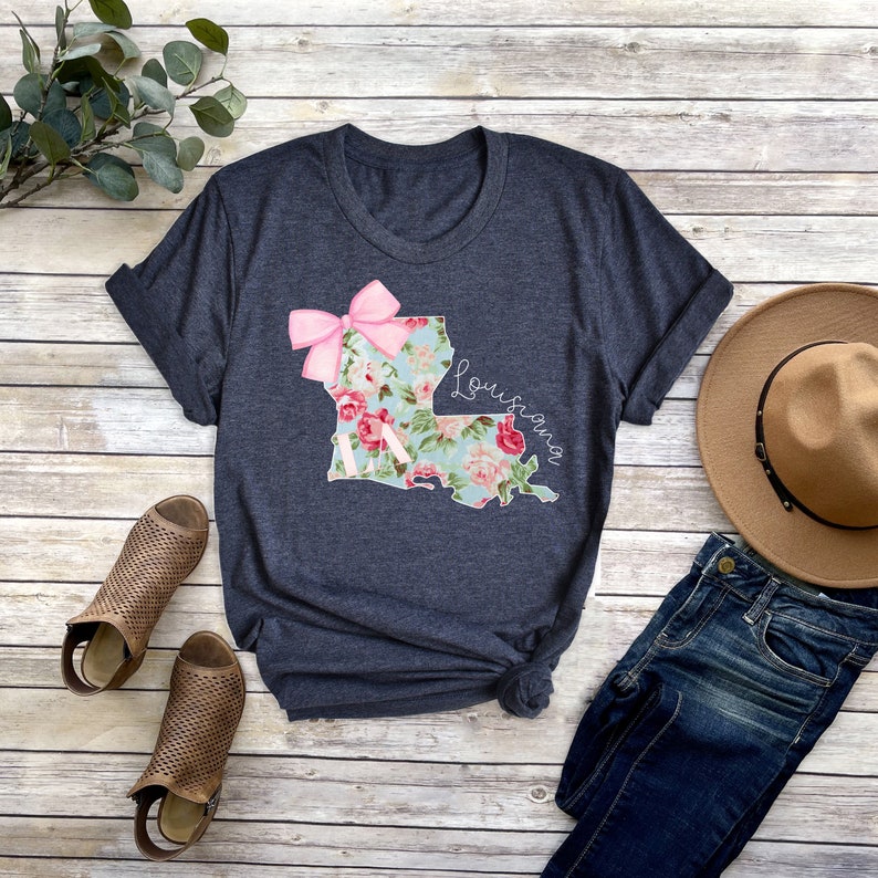 May include: A dark blue t-shirt with a floral print of the state of Louisiana. The print includes a pink bow and the word "Louisiana".