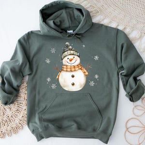 May include: A green hooded sweatshirt featuring a watercolor snowman design. The snowman wears a black and gray hat, an orange and brown scarf, and has a carrot nose. White snowflakes are scattered around the snowman.