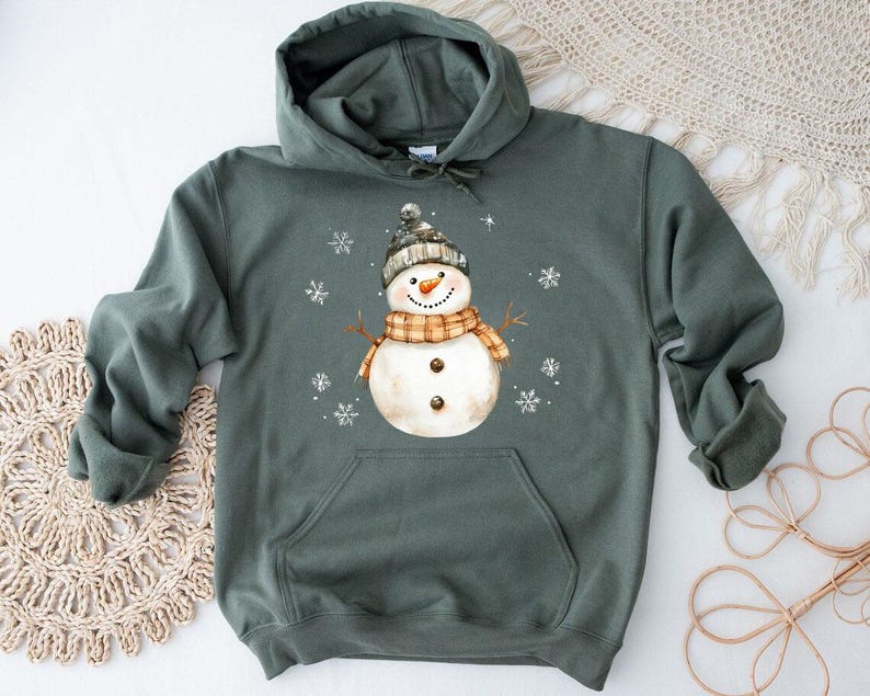 May include: A green hooded sweatshirt featuring a watercolor snowman design. The snowman wears a black and gray hat, an orange and brown scarf, and has a carrot nose. White snowflakes are scattered around the snowman.