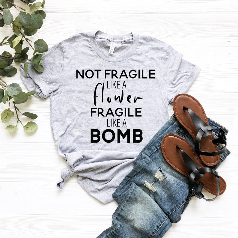 May include: A heather gray t-shirt with the text "NOT FRAGILE LIKE A flower FRAGILE LIKE A BOMB" in black. The shirt is paired with blue denim jeans and black sandals with brown soles.