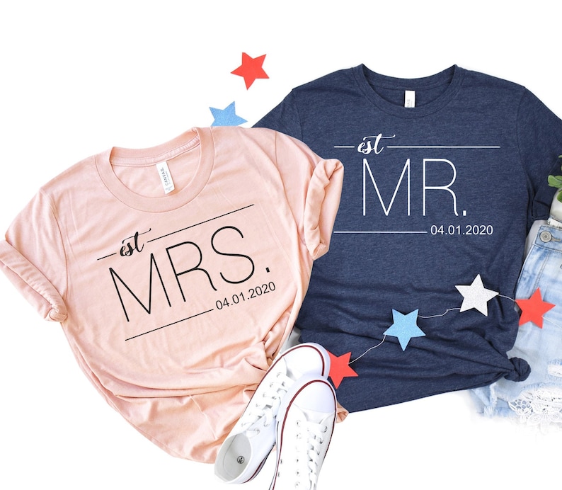 May include: Two t-shirts, one pink and one navy blue, with the text "est. MRS. 04.01.2020" and "est. MR. 04.01.2020" printed on them. The shirts are laid out on a white background with red, white, and blue stars scattered around them.