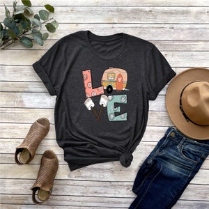 May include: A dark gray t-shirt with a graphic of the word "LOVE" with a camper van inside the "O". The letters are decorated with swirls and dots. There are two marshmallows on sticks next to the "E".