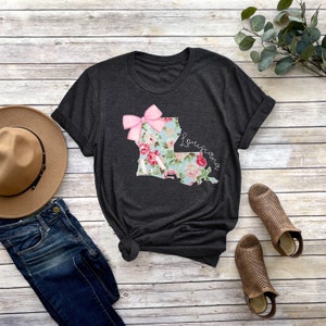 May include: A dark grey t-shirt with a floral print of the state of Louisiana with a pink bow on top. The text "Louisiana" is written in cursive on the right side of the state outline.