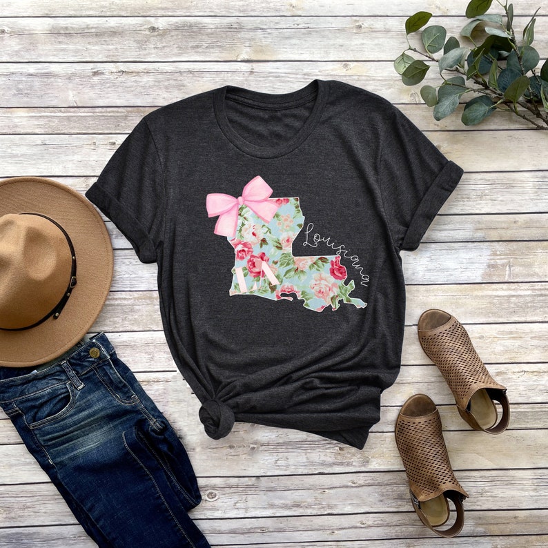 May include: A dark grey t-shirt with a floral print of the state of Louisiana with a pink bow on top. The text "Louisiana" is written in cursive on the right side of the state outline.